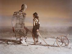 two people standing next to each other in front of a skeleton on the beach with bicycles