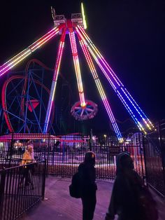 people walking around an amusement park at night with lights on the ferris wheel in the background