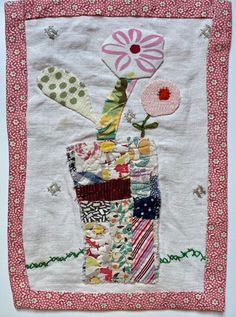 a patchwork quilted wall hanging with a flower in a vase on the side