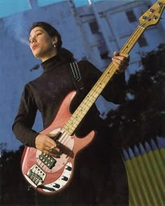 a woman holding a pink bass guitar in front of a blue sky and white building