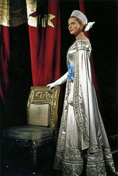 a woman in an elaborate dress and tiara standing next to a chair with red curtains behind her