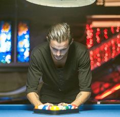 a man in black shirt playing pool with colorful balls on blue table and lights behind him