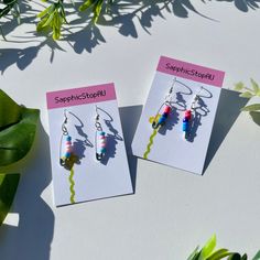 two earrings with colorful beads hanging from them on top of a white table next to green leaves