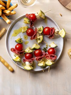 a white plate topped with cucumber and tomato skewers next to bread sticks