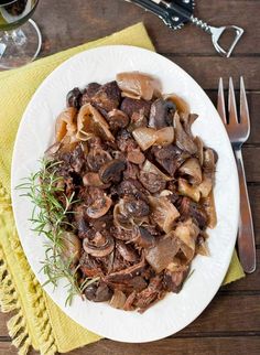 a white plate topped with meat and mushrooms next to a fork, knife and glass of wine