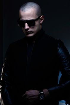 a man in black jacket and sunglasses standing with his hand on his hip while looking at the camera