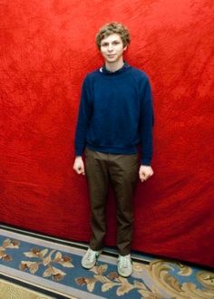 a young man standing in front of a red wall with his hands in his pockets