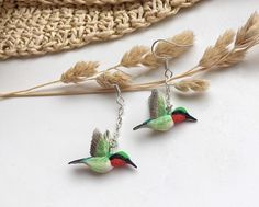 two hummingbirds are hanging from silver earwires on a white surface next to some dried grass