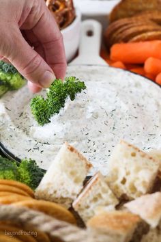 a person dipping broccoli into a bowl of ranch dip with crackers and carrots