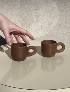 two coffee mugs on a table being held by someone's hand with their fingers