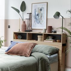 a bed with green sheets and pillows next to a book shelf filled with plants in front of a pink wall