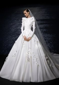 a woman in a white wedding dress with flowers on the skirt and veil, standing against a dark background