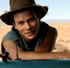 a young man wearing a hat leaning on the hood of a blue car in an arid area