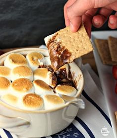 someone dipping marshmallows into a bowl of hot chocolate and graham crackers