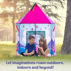 two children sitting in front of a play tent on the grass with trees behind them