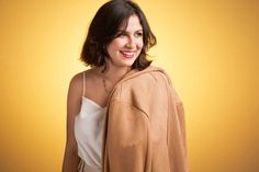 a woman in a white top and tan jacket smiling at the camera while standing against a yellow background