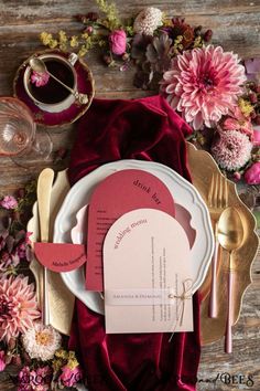 a place setting with pink flowers and goldware on a wooden tablecloth, along with red velvet napkins