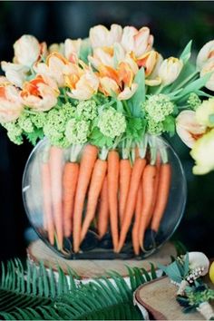 carrots and flowers in a glass vase on a table with greenery next to it