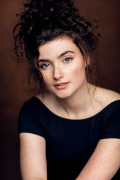 a woman with curly hair sitting in front of a brown background wearing a black shirt