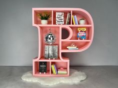 a pink letter shaped shelf with books and other items on it's sides, in front of a gray wall