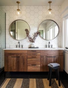 two round mirrors are above the double sinks in this bathroom with wood cabinetry and white hexagonal tiles