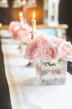 pink roses are in a square vase on a table with white linens and candles