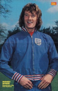 a man with long hair wearing a blue jacket and striped pants, standing in front of a green field