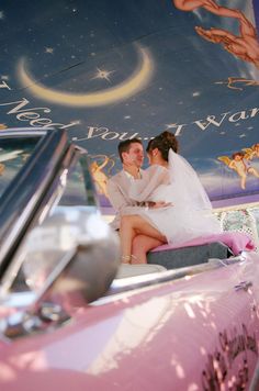 a bride and groom sitting on top of a pink car in front of a mural