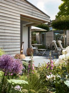 a garden with flowers and chairs in front of a wooden building that has an open patio area