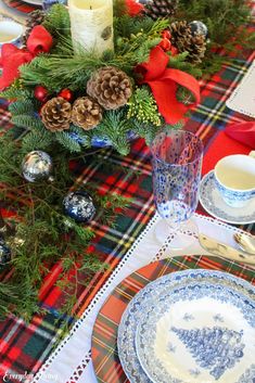 the table is set for christmas dinner with blue and white plates, silverware, pine cones, red plaid napkins