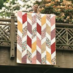 a woman holding up a quilt on top of a wooden bridge next to flowers and trees