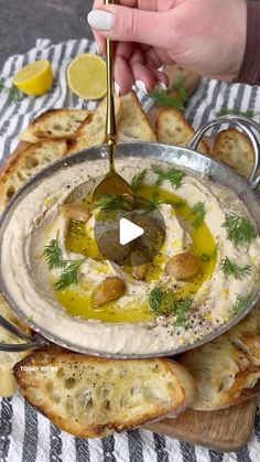 a person spooning olives into a bowl of hummus with garlic bread on the side