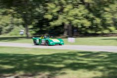 a green race car driving down a road in the grass with trees and signs behind it