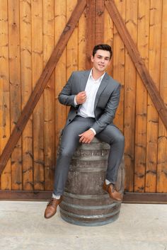 a man sitting on top of a barrel in front of a wooden wall wearing a suit