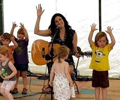 a woman singing into a microphone while children play with guitar behind her and on the ground