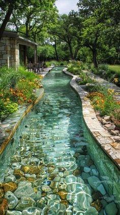 a river that is surrounded by flowers and rocks in the water, next to a house