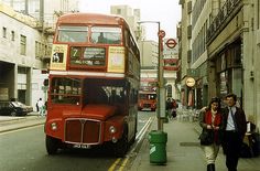 two people walking on the sidewalk next to a red double decker bus in an urban area