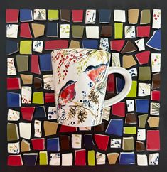 a coffee cup sitting on top of a colorful mosaic tile covered wall with red, white and blue birds