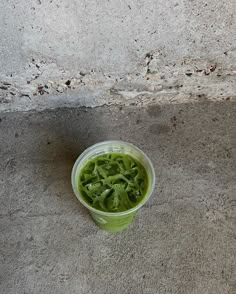 a cup filled with green liquid sitting on top of a cement floor next to a wall