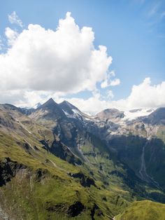 the mountains are covered with green grass and snow in the distance, under a partly cloudy blue sky