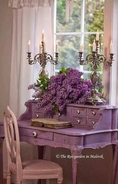 a purple desk with flowers and candles on it in front of a window, next to a chair