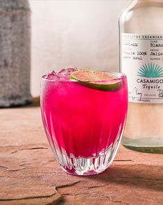 a pink drink with a slice of lime on the rim and a bottle behind it