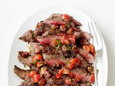 a white plate topped with sliced steak and tomato salad next to a fork on top of a table