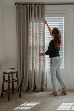 a woman standing in front of a window opening curtains