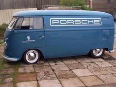 an old blue van parked on top of a brick road next to a building with a sign that reads porsche