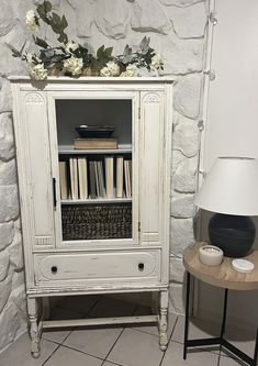 a white cabinet sitting next to a table with a lamp on it and flowers in the corner