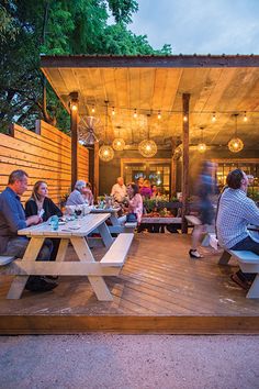 people are sitting at picnic tables outside in the evening time, with lights hanging above them