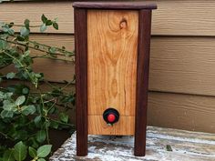 a wooden box with a red button on it sitting in front of a house next to a bush