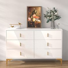 a white dresser with flowers on top and a painting above it in a room that has hardwood floors