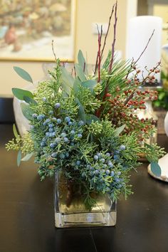 a vase filled with blue berries and greenery on top of a table next to a candle